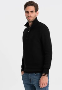 Maglione nero a maglia con colletto alto e mezza zip. Caratterizzato da un tessuto testurizzato e polsini a coste, abbinato a jeans blu e un bracciale.