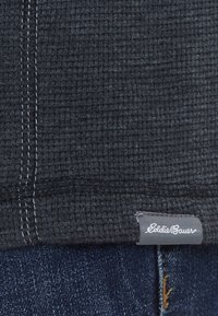 Tissu gris foncé texturé avec double couture verticale à côté d'une petite étiquette en tissu Eddie Bauer au-dessus d'un jean en denim bleu.