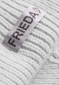 Witte gebreide stof met een textuur, ribbelpatroon. Een lichtpaarse label toont de tekst "Friedag" in donkergrijs.