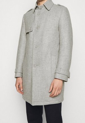 Homme portant un manteau en laine gris clair boutonné avec des sangles à boucle aux poignets et un pantalon noir, sur un fond uni.