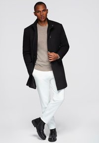 Zwarte jas met hoge kraag over een beige trui, witte broek en zwarte laarzen. Glad stof met minimale hardwaredetails.