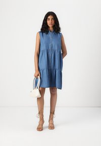 Τζιν φόρεμα χωρίς μανίκια με κουμπωτό μπροστά, σε tiered σχέδιο και χαλαρή εφαρμογή. Συνδυάζεται με λευκή τσάντα και καφέ δερμάτινες σανδάλια με κορδόνια.