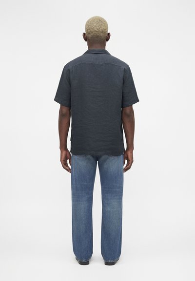 Άνδρας με κοντά ξανθά μαλλιά, φοράει μαύρο κοντομάνικο πουκάμισο και μπλε τζιν, στέκεται γυρισμένος με την πλάτη σε λευκό φόντο.