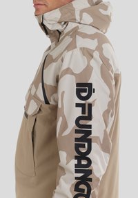 Tarnjacke in Tan und Hellbeige. Verfügt über eine Kapuze, Fronttasche und schwarzen Logo-Text auf dem Ärmel. Glattes, wetterbeständiges Material.