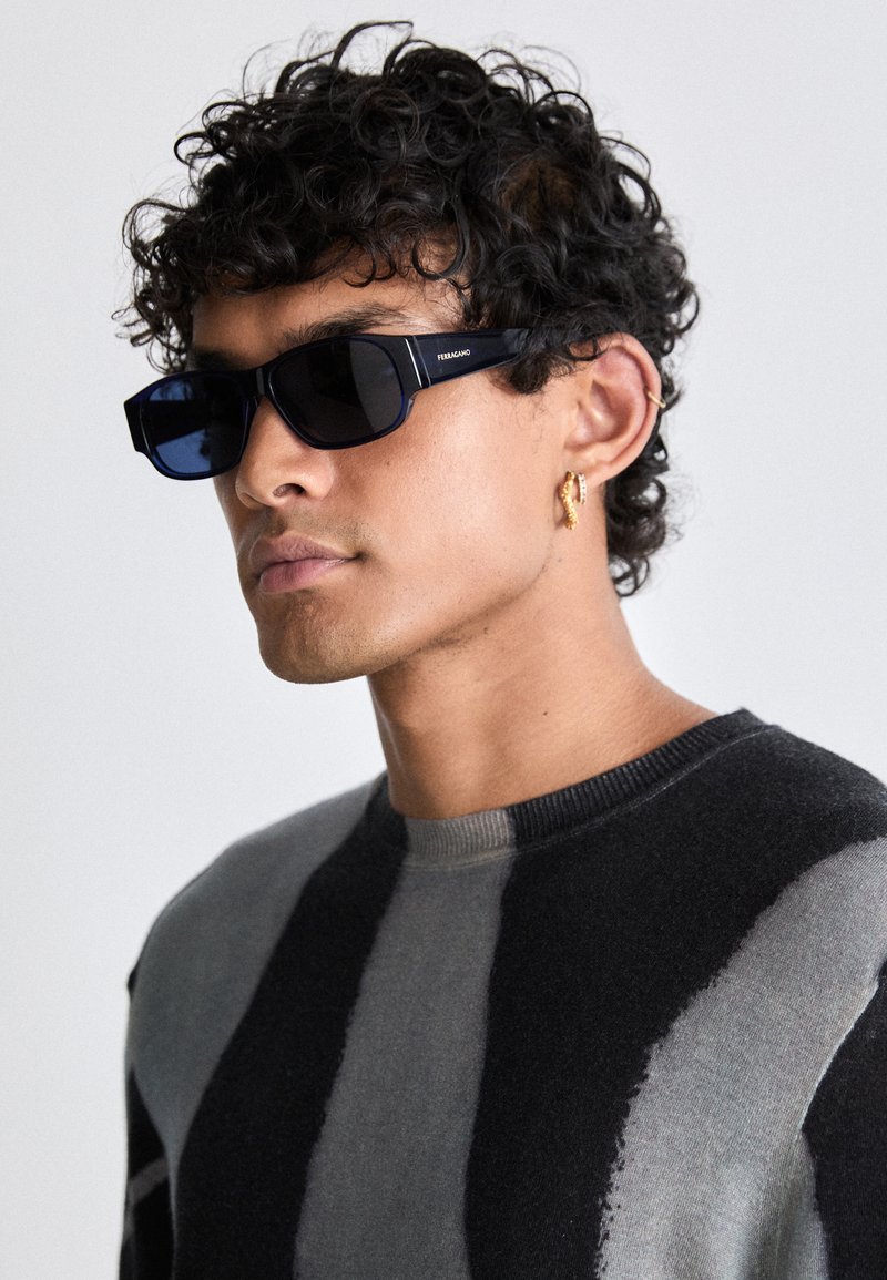 Uomo con capelli ricci che indossa occhiali da sole neri rettangolari, orecchini d'oro e un maglione a strisce nere e grigie, rivolto verso sinistra.