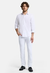 Chemise blanche à manches longues et boutonnée avec une coupe droite et des manches retroussées, associée à un pantalon blanc et des chaussures noires à lacets.