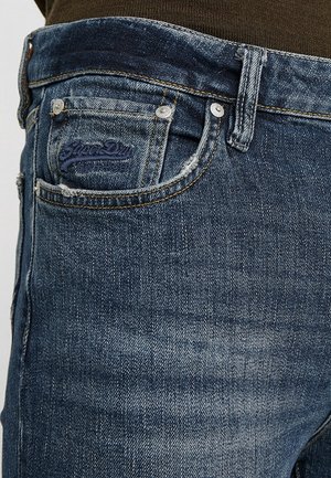 Close-up af blå denimjeans med falmet tekstur, lille frontlomme med metalnitter og mørkt broderet brandlogo på lommen.