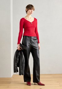 Haut long à manches longues rouge côtelé avec un col zippé, associé à un pantalon large en cuir noir et des escarpins rouges à bout pointu, tenant une veste en cuir noire.