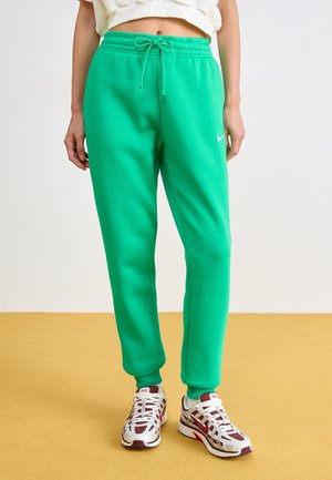 Personne portant un pantalon de survêtement vert Nike avec cordon et des baskets Nike blanches et rouges, debout sur un sol jaune.