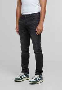 Man in zwarte distressed skinny jeans, een wit tanktop en witte en groene Nike sneakers staand op een effen achtergrond.