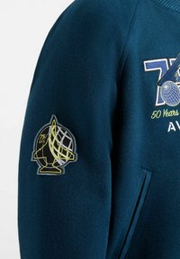 Donkerblauwe jas mouw met een geel en zwart ruimtehelm patch en gedeeltelijke geborduurde tekst met de woorden "70," "50 Jaar," en "AVX."