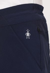 Tmavě modré šortky s diagonálním švem. Na levé straně mají malé reflexní logo postavičky a bezpečné kapsy na zip.