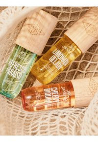 Tres coloridas botellas de spray de perfume Sol de Janeiro con etiquetas Refresco Paraíso, Limonada Gelada y Cheeky Biquini, con tapas de madera sobre tela de encaje.