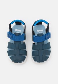 Mėlyna šlepetė su nertine viršutine dalimi, reguliuojama sagtimi ir paminkštintu įdėklu. Yra balta gumine padais. Prekės ženklas "Camper" matomas ant įdėklo.