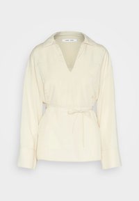 Blouse crème avec un décolleté en V, des manches longues et une taille nouée. Fabriquée en tissu doux avec une texture lisse et une coupe décontractée.