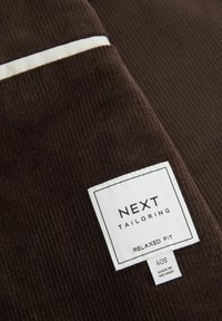 Hnědá kordurová látka s ribbovanou texturou; obsahuje bílou etiketu označenou "NEXT TAILORING RELAXED FIT," která uvádí velikost 40S a zemi původu.