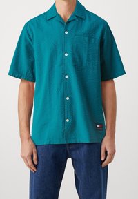 Camicia a maniche corte color turchese con un motivo testurizzato, chiusura con bottoni e taschino sul petto. Presenta un'etichetta con logo sull'orlo.