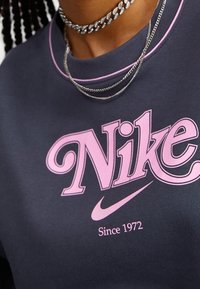 Sudadera de Nike en gris oscuro con logo y texto rosa "Desde 1972." Cuenta con un cuello redondo y cadenas plateadas superpuestas. Textura suave.
