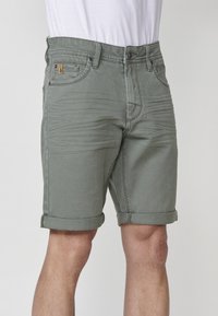 Shorts en denim vert avec un ourlet roulé, comportant des poches avant, une texture subtile et un patch de marque sur la taille.