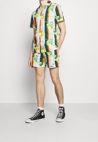 Set van wit T-shirt met korte mouwen en korte broek met patronen van tropisch fruit en bladeren in groen, oranje en geel, gecombineerd met zwarte hoge sneakers.