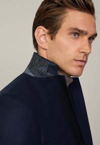 Cappotto navy con colletto alto e dettagli in lana grigia. Il design è elegante, con una forma strutturata e una texture liscia.