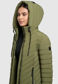 Giacca imbottita verde oliva con cappuccio, caratterizzata da una trama a cuciture diagonali, zip nere e cordini regolabili per una vestibilità ottimale.