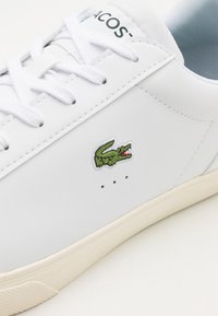 Baskets en cuir blanc avec des détails cousus, logo crocodile vert brodé, et semelle extérieure en caoutchouc de couleur claire. Chaussures à lacets plats blancs.
