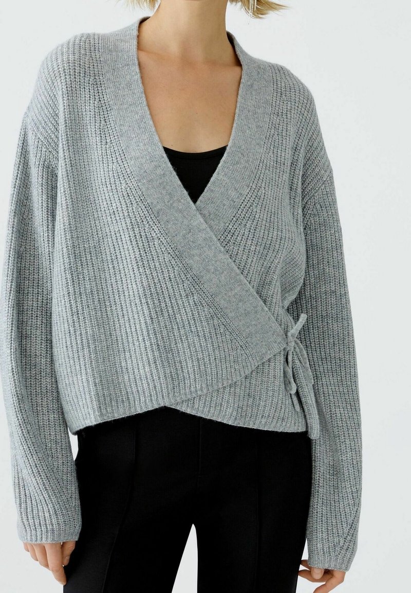 Pull gris côtelé enveloppant avec un col en V profond, des manches amples et une fermeture à nouer sur le côté. Porté sur un haut noir et associé à un pantalon noir.