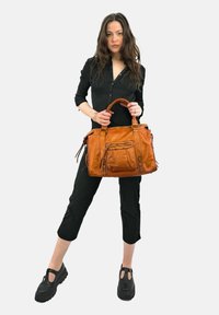 Femme en tenue noire tenant un grand sac en cuir beige avec deux poignées, se tenant devant un fond clair.
