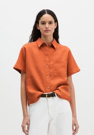 Kobieta o długich ciemnych włosach, nosząca pomarańczową koszulę na krótki rękaw wpuszczoną w białe spodnie z czarnym paskiem.