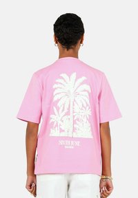 Roze t-shirt met korte mouwen, met een witte palmbomenafbeelding op de achterkant en de tekst "SIXTH JUNE PARADISE" nabij de zoom.