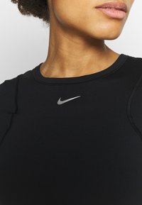 Top atlético negro de Nike con cuello redondo, mangas ajustadas y un detalle de logo plateado en el pecho. Tela suave y elástica con costuras.