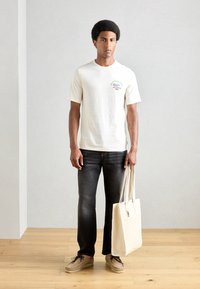 Ljusbeige bomullströja med korta ärmar, mörk denimjeans, beige mockaskor och en krämfärgad toteväska. Trägolv som bakgrund.