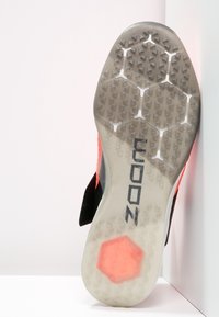 La suola della scarpa presenta un motivo esagonale con piccole impugnature triangolari, di colore grigio chiaro, e un lato in tessuto nero con un'accensione arancione. Il testo recita "COON."