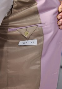 Brun og rosa tekstilinnredning av et plagg med en knappet lomme. En etikett med teksten "PIER•ONE" er synlig over lommen.