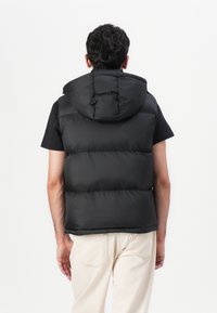 Melnā pufīgā veste ar kapuci, ar quilted dizainu un brīvu piegriezumu. Valkāta virs melna īsām piedurknēm krekliem un gaišu biksēm.