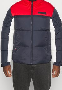 Pufová bunda s vysokým límcem. Červená horní část, námořnický modrý trup a bílé rukávy. Obsahuje přední zip a boční kapsy. Měkká, prošívaná textura.