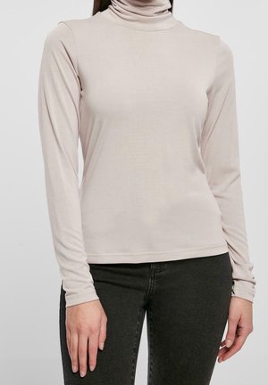 Beige coltrui met lange mouwen gedragen met zwarte broek, afgebeeld van schouders tot heupen tegen een effen achtergrond.