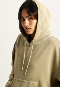 Przesadny, beżowy sweatshirt z kapturem wykonany z mieszanki bawełny, z kieszenią z przodu oraz regulowanymi sznurkami zakończonymi srebrnymi końcówkami. Dyskretny, haftowany logo.