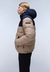 Giacca imbottita beige e navy con cappuccio, dotata di chiusura con zip, tasche laterali e dettaglio del logo sulla spalla. Texture liscia.