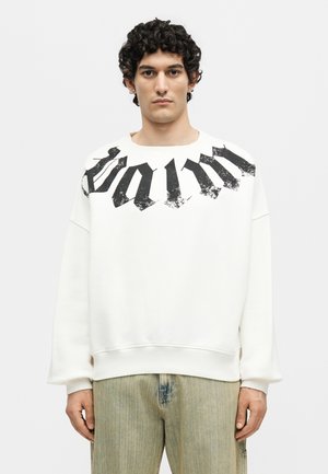 Νεαρός άνδρας με κατσαρά μαλλιά, φορώντας λευκό φούτερ με μεγάλο μαύρο γοτθικό κείμενο στο στήθος και ανοιχτό πράσινα τζιν.