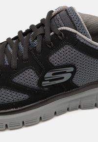 Sapatilha atlética em preto e cinza com parte superior em malha texturizada, colarinho acolchoado, atacadores pretos e sola em borracha cinza com um padrão de superfície distinto.