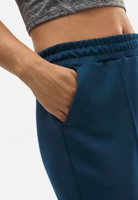 Pantalones deportivos azules con cintura elástica y bolsillo lateral; la tela parece suave y lisa, diseñada para la comodidad y la funcionalidad.