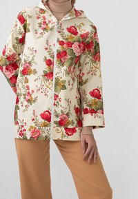 Mujer vistiendo una sudadera cremosa con cremallera y estampado floral en rojo, rosa y amarillo, combinada con pantalones beige y un suéter de cuello alto color beige.