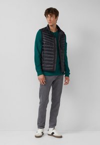 Jonge man draagt een zwarte puffer vest over een groene lange mouwen shirt, grijze jeans en witte sneakers, staande tegen een effen witte achtergrond.