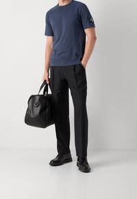 Mies, joka käyttää tummansinistä lyhythihaista paitaa ja mustia housuja, pitää käsissään mustaa nahkareppua, seisoen tasaisella vaalealla taustalla.