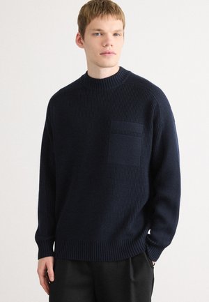 Navy blue knitted sweater with a high neck and textured fabric. Features a front pocket and ribbed cuffs and hem.