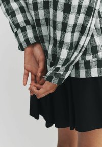 Bluza verde și albă cu model în carouri, mâneci lungi, manșete cu nasturi și țesătură texturată. Fustă neagră vizibilă în partea de jos, cu un design evazat.