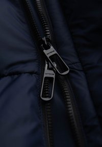 Czarne zamki błyskawiczne na ciemnogranatowej puchowej kurtce, z pikowaną teksturą i firmowymi uchwytami. Zbliżenie uwydatnia detale szycia.