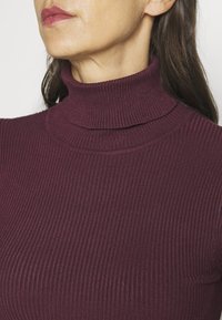 Burgundowy sweter z golfem o prążkowanej fakturze, dopasowany krój i warstwowy detal przy szyi, wykonany z elastycznego materiału, przedstawiony od pasa w górę.